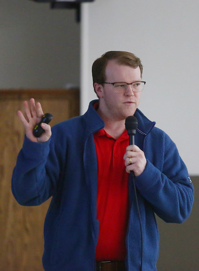 Man speaking with one hand raised, other holding a microphone