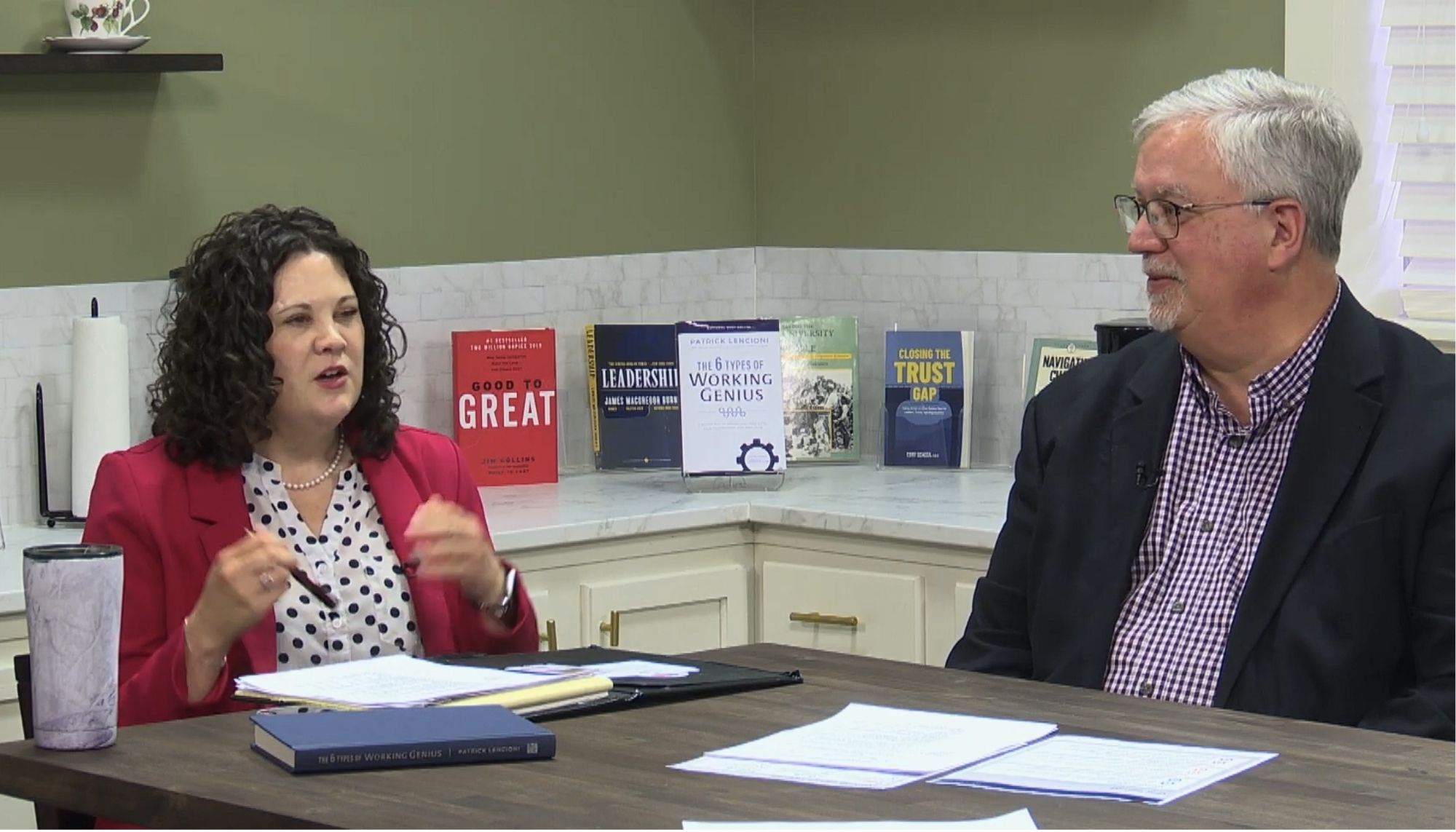 A white woman and white man are sitting at a table with books and papers surrounding them